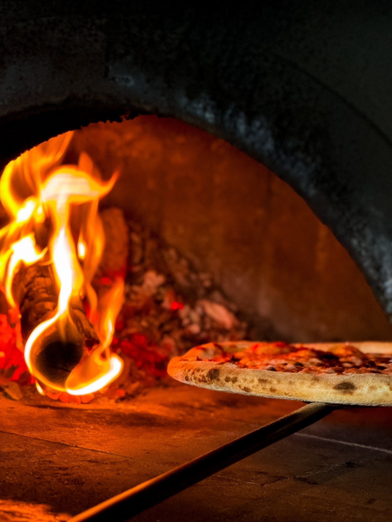 Pizza on a peel beside the flames inside the wood-fired oven